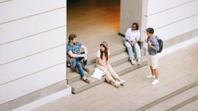 Vier internationale Studierende sitzen und stehend auf den Stufen zum Eingangsbereich einer Universität.